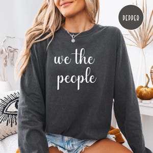 Puede incluir: Una sudadera gris oscuro de manga larga y cuello redondo. La sudadera tiene el texto "We the People" impreso en blanco en la parte delantera.
