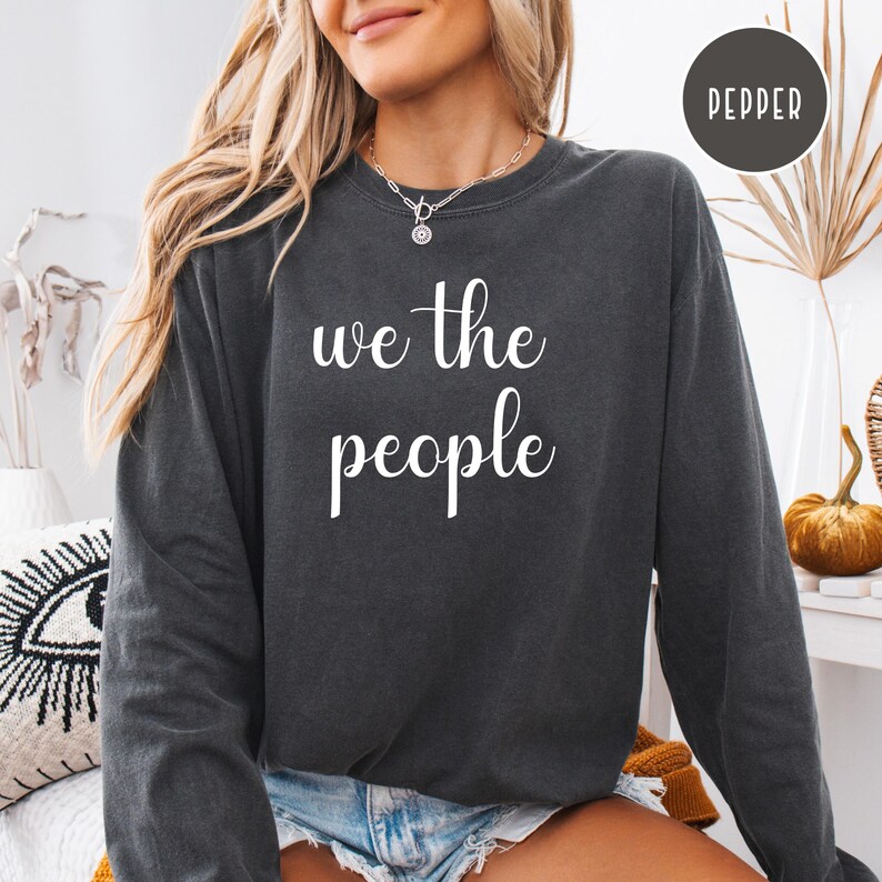 Puede incluir: Una sudadera gris oscuro de manga larga y cuello redondo. La sudadera tiene el texto "We the People" impreso en blanco en la parte delantera.