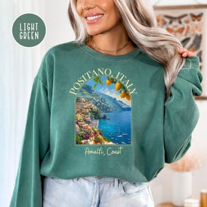 K&ouml;nnte beinhalten: Ein hellgr&uuml;nes Sweatshirt mit einem Grafikdruck von Positano, Italien, mit einer farbenfrohen Stadtlandschaft, einem blauen Meer und Zitronen an einem Baum. Der Text "Positano, Italien" und "Amalfik&uuml;ste" ist auf dem Sweatshirt aufgedruckt.