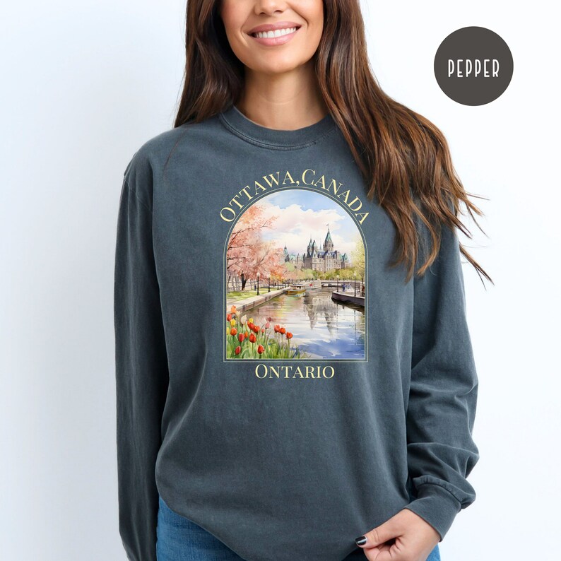 May include: A dark gray long-sleeve sweatshirt with a graphic print of a watercolor illustration of the Parliament Buildings in Ottawa, Canada. The text "OTTAWA, CANADA" is printed in a yellow arch above the illustration. The text "ONTARIO" is printed in yellow below the illustration.