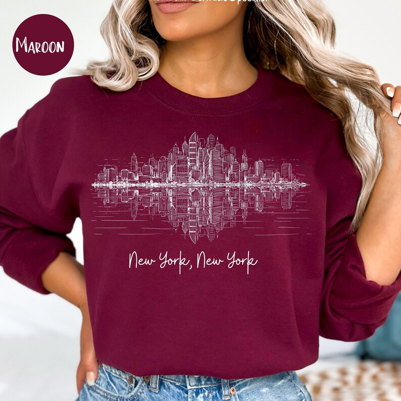 K&ouml;nnte beinhalten: Ein maroonfarbener Pullover mit einem wei&szlig;en Grafikdruck des New Yorker Stadtbildes, das sich im Wasser spiegelt. Der Text "New York, New York" ist unter dem Grafikdruck aufgedruckt.