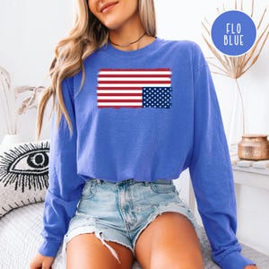 Puede incluir: Una camiseta azul de manga larga con un diseño de bandera estadounidense desgastada. El diseño de la bandera incluye rayas rojas y blancas y un campo azul de estrellas. Un logotipo circular con el texto "FLO BLUE" es visible.