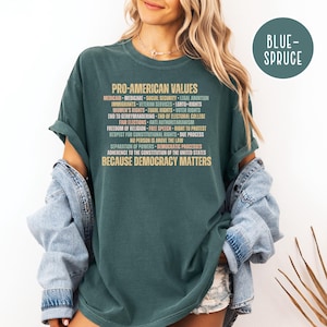 Pode incluir: T-shirt verde escuro com o texto "PRO-AMERICAN VALUES" e uma lista de valores. A camisa tamb&eacute;m tem o texto "BECAUSE DEMOCRACY MATTERS". Um c&iacute;rculo azul-esverdeado com o texto "BLUE-SPRUCE" est&aacute; no canto superior direito.