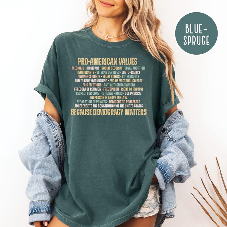 Pode incluir: T-shirt verde escuro com o texto "PRO-AMERICAN VALUES" e uma lista de valores. A camisa tamb&eacute;m tem o texto "BECAUSE DEMOCRACY MATTERS". Um c&iacute;rculo azul-esverdeado com o texto "BLUE-SPRUCE" est&aacute; no canto superior direito.