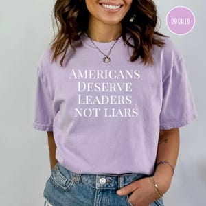 May include: A light purple t-shirt with the text "Americans Deserve Leaders Not Liars" printed in white.