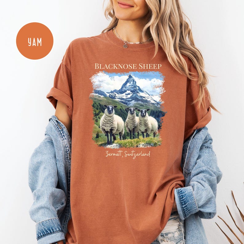 K&ouml;nnte beinhalten: Rostfarbenes T-Shirt mit einer Grafik von drei Schwarznasenschafen vor einer Bergkette. Der Text "BLACKNOSE SHEEP" und "Zermatt, Switzerland" sind oberhalb und unterhalb des Bildes aufgedruckt. Das Shirt hat kurze &Auml;rmel.
