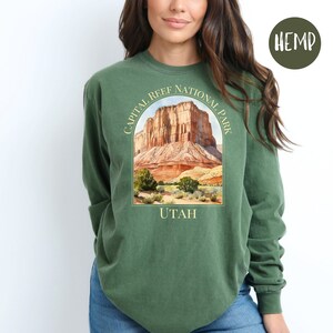 May include: Green long-sleeve shirt with a graphic of Capital Reef National Park in Utah. The graphic features a watercolor illustration of the park's rock formations and the text "CAPITAL REEF NATIONAL PARK" and "UTAH". The shirt has a "HEMP" label.