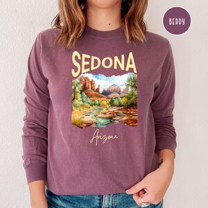 May include: A purple long-sleeved sweatshirt with a graphic print of a landscape scene with red rock formations and a river. The text "SEDONA Arizona" is printed on the shirt.