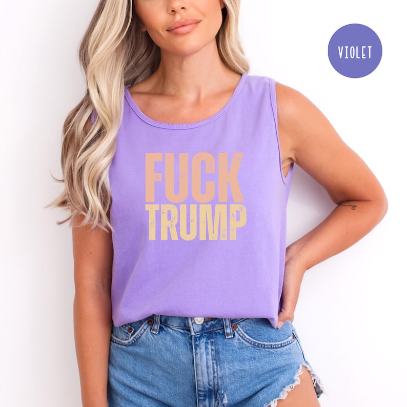 K&ouml;nnte beinhalten: Violettes Tanktop mit dem Text "FUCK TRUMP" in einer verblassten, hellorangen Schriftart. Das Tanktop ist &auml;rmellos. Ein violetter Kreis mit dem Wort "VIOLET" befindet sich in der oberen rechten Ecke. Das Oberteil wird mit blauen Jeansshorts getragen.