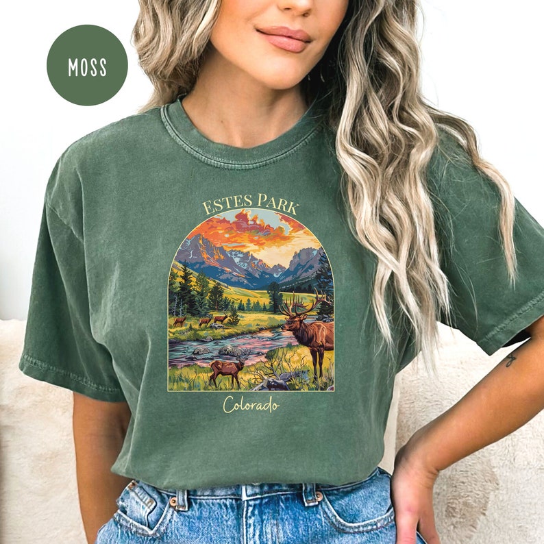May include: A green t-shirt featuring a vintage-style illustration of a mountain landscape with an elk in the foreground. The text "ESTES PARK Colorado" is printed on the shirt.