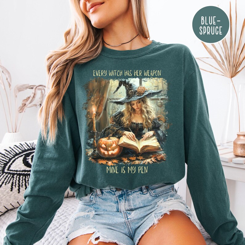 May include: Teal long-sleeve sweatshirt featuring a witch illustration with the text "EVERY WITCH HAS HER WEAPON, MINE IS MY PEN." The graphic depicts a witch writing in a book with a pumpkin and candle. The sweatshirt is a comfortable, casual style.
