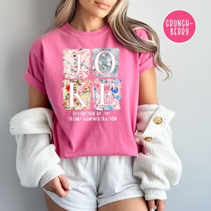 May include: Pink t-shirt with a floral design spelling out the word "JOKE" and the text "Definition of the Trump Administration".