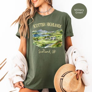 Puede incluir: Camiseta verde militar con una ilustraci&oacute;n en acuarela de las Tierras Altas escocesas. El dise&ntilde;o incluye un paisaje con monta&ntilde;as, un lago y edificios, con el texto "SCOTTISH HIGHLANDS" y "Scotland, UK".