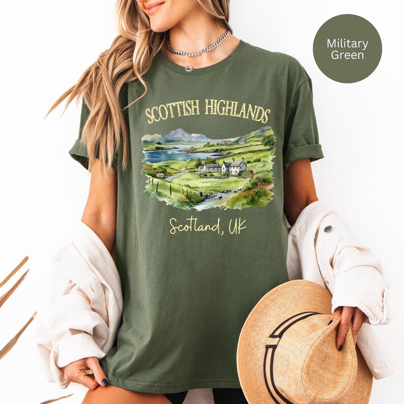 Puede incluir: Camiseta verde militar con una ilustraci&oacute;n en acuarela de las Tierras Altas escocesas. El dise&ntilde;o incluye un paisaje con monta&ntilde;as, un lago y edificios, con el texto "SCOTTISH HIGHLANDS" y "Scotland, UK".