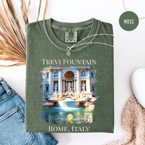 May include: A moss green t-shirt with a graphic of the Trevi Fountain in Rome, Italy. The shirt has a crew neck and short sleeves. A silver necklace with a sun pendant is draped across the shirt.