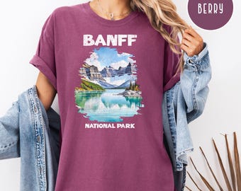 May include: Berry-colored t-shirt with a graphic of Banff National Park. The design features a mountain range, a lake, and the text "BANFF NATIONAL PARK" in white. The shirt has rolled-up sleeves.