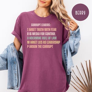 May include: Berry-colored t-shirt with the text "CORRUPT LEADERS: TARGET TRUTH WITH FEAR, RIG MEDIA FOR CONTROL, UNDERMINE RULE OF LAW, MARKET LIES AS LEADERSHIP, PARDON THE CORRUPT." The shirt has rolled-up sleeves and a relaxed fit. A circular "BERRY" label is in the upper right corner.
