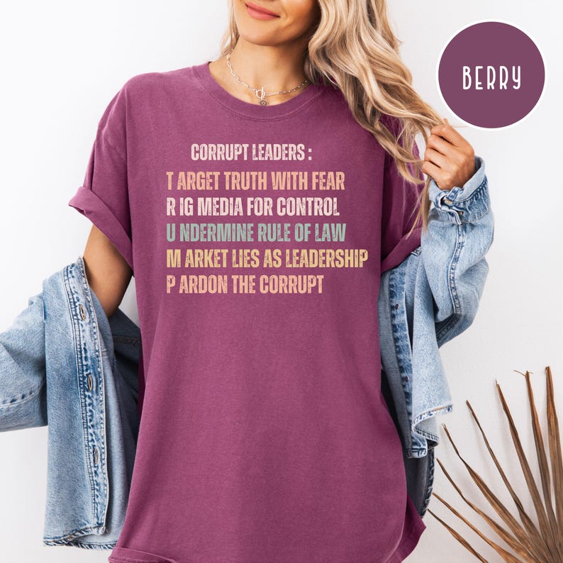 May include: Berry-colored t-shirt with the text "CORRUPT LEADERS: TARGET TRUTH WITH FEAR, RIG MEDIA FOR CONTROL, UNDERMINE RULE OF LAW, MARKET LIES AS LEADERSHIP, PARDON THE CORRUPT." The shirt has rolled-up sleeves and a relaxed fit. A circular "BERRY" label is in the upper right corner.