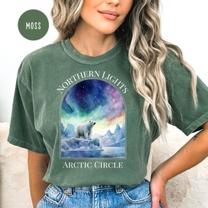 Op de afbeelding: Mosgroen T-shirt met een afbeelding van een ijsbeer op een ijsberg onder het noorderlicht. De tekst "NORTHERN LIGHTS" en "ARCTIC CIRCLE" staat erop. Het T-shirt heeft een relaxte pasvorm.
