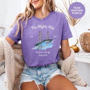 May include: A purple t-shirt with the text "The Mighty Mac" and an illustration of the Mackinac Bridge. The shirt also includes the text "Est. 1957" and "Mackinac Bridge Walk 2025". The shirt is a heather purple color.