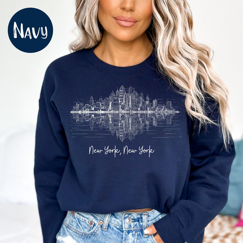 K&ouml;nnte beinhalten: Ein marineblaues Sweatshirt mit einer wei&szlig;en Grafik der Skyline von New York City, die im Wasser reflektiert wird. Der Text "New York, New York" ist unter der Grafik aufgedruckt.