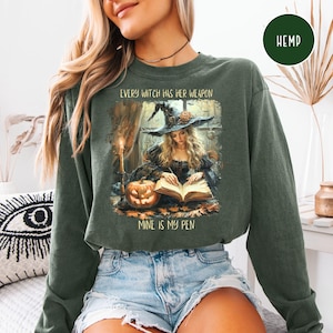 May include: Dark green sweatshirt with the text "EVERY WITCH HAS HER WEAPON, MINE IS MY PEN." The graphic depicts a witch writing in a book, with a pumpkin and candle. The sweatshirt has long sleeves and a crew neck.