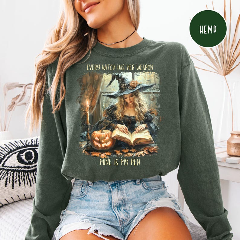 May include: Dark green sweatshirt with the text "EVERY WITCH HAS HER WEAPON, MINE IS MY PEN." The graphic depicts a witch writing in a book, with a pumpkin and candle. The sweatshirt has long sleeves and a crew neck.