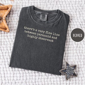 May include: Dark gray t-shirt with the text "there's a very fine line between paranoid and highly observant" in white. The shirt is folded and lies on a white textured surface. A small star-shaped decoration is visible.