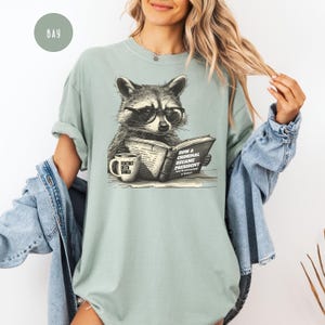 May include: A light green t-shirt featuring a raccoon wearing glasses and reading a book titled "How a Criminal Became President." A coffee mug with the text "Foxtrot Delta Tango" sits beside the book. The shirt has a relaxed fit.