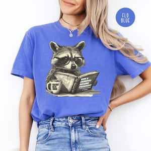 May include: Blue t-shirt featuring a raccoon wearing glasses, reading a book titled "How a Criminal Became President." A coffee mug with "Foxtrot Delta Tango" is next to the book. The shirt has a crew neck and short sleeves.
