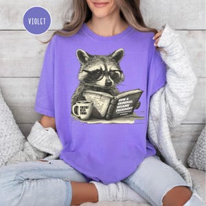 May include: Violet t-shirt featuring a raccoon wearing glasses and reading a book titled "How a Criminal Became President." A coffee mug with the text "Foxtrot Delta Tango" is also visible. The shirt is a comfortable, casual style.