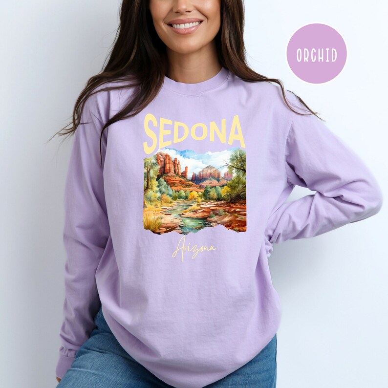 May include: A lavender long-sleeve t-shirt with a graphic print of a landscape scene in Sedona, Arizona. The text "Sedona" is printed in yellow above the image and "Arizona" is printed in yellow below the image.