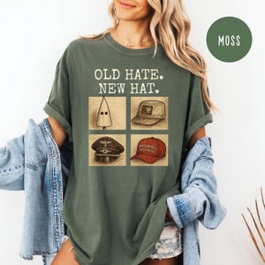 Könnte beinhalten: Moosgrünes T-Shirt mit dem Text "OLD HATE. NEW HAT." und vier Hutillustrationen. Die Illustrationen zeigen eine weiße, spitze Kapuze, eine beige Baseballkappe, eine schwarze Militärmütze und eine rote "MAKE AMERICA GREAT AGAIN"-Kappe.