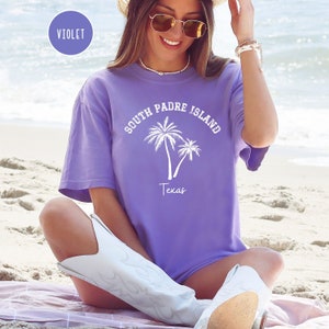 May include: A purple t-shirt with a white graphic of palm trees and the text "South Padre Island Texas".