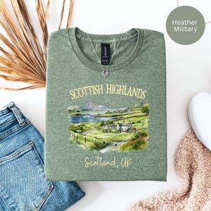 Puede incluir: Camiseta verde militar jaspeada con un dise&ntilde;o de acuarela de las Tierras Altas escocesas. El dise&ntilde;o incluye un paisaje con un lago, casas y el texto "Scottish Highlands" y "Scotland, UK" en letras doradas. Se ve un collar plateado.