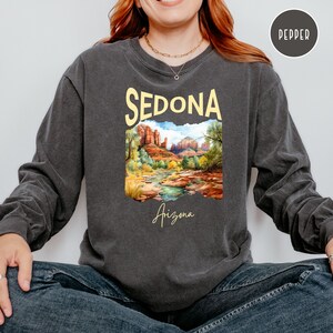 May include: A charcoal grey long-sleeve shirt with a vintage-style graphic of a landscape in Sedona, Arizona. The graphic features red rock formations, a river, and trees. The text "Sedona" is in yellow and the text "Arizona" is in a cursive font.