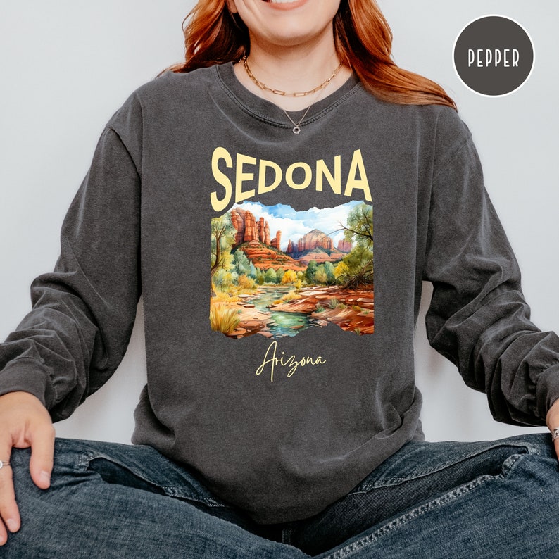 May include: A charcoal grey long-sleeve shirt with a vintage-style graphic of a landscape in Sedona, Arizona. The graphic features red rock formations, a river, and trees. The text "Sedona" is in yellow and the text "Arizona" is in a cursive font.