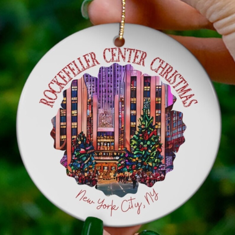 May include: A white ceramic ornament with a colorful image of the Rockefeller Center Christmas tree and the surrounding buildings. The text "Rockefeller Center Christmas New York City, NY" is written in red on the ornament.