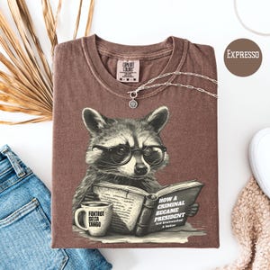 May include: A brown Comfort Colors t-shirt featuring a raccoon wearing glasses and reading a book titled "How a Criminal Became President". A coffee mug with "Foxtrot Delta Tango" is next to the book. The shirt is folded with a silver necklace.