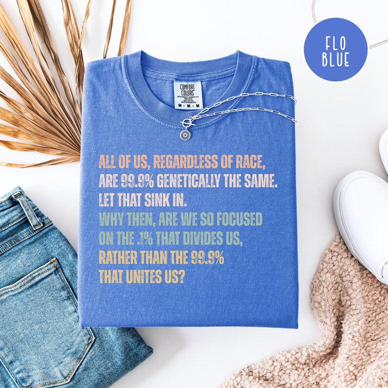 May include: A blue Comfort Colors t-shirt with a silver necklace. The shirt features text in multiple colors: "ALL OF US, REGARDLESS OF RACE, ARE 99.9% GENETICALLY THE SAME. LET THAT SINK IN. WHY THEN, ARE WE SO FOCUSED ON THE .1% THAT DIVIDES US, RATHER THAN THE 99.9% THAT UNITES US?"