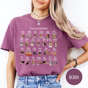 May include: A purple t-shirt with a graphic of a dog alphabet. Each letter of the alphabet is represented by a dog breed with a corresponding word describing the breed. The text "The Dog Alphabet" is at the top of the graphic.