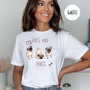 Puede incluir: Camiseta blanca con un gr&aacute;fico de tres carlinos y el texto "Hugs and Pugs" en morado.