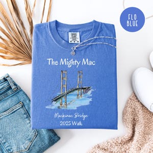 May include: A blue t-shirt with the text "The Mighty Mac" and an illustration of the Mackinac Bridge. The shirt also has the text "Mackinac Bridge 2025 Walk". A silver necklace with a pendant is on the shirt.