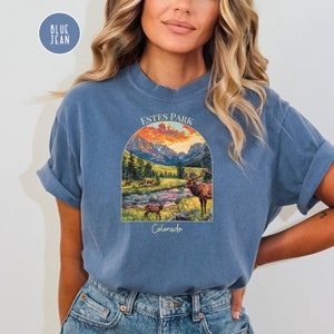 May include: A blue jean t-shirt featuring a graphic of a mountain scene with a sunset and elk. The text "Estes Park Colorado" is printed on the shirt.