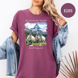 K&ouml;nnte beinhalten: Beerenfarbenes T-Shirt mit einer Grafik von Schwarznasenschafen vor einer Bergkette. Der Text "BLACKNOSE SHEEP" und "Zermatt, Switzerland" sind auf dem Shirt aufgedruckt. Die &Auml;rmel sind hochgekrempelt.