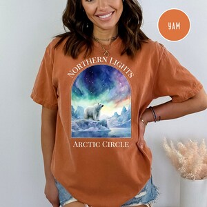Op de afbeelding: Een roestkleurig t-shirt met een afbeelding van een ijsbeer op een ijsberg onder het noorderlicht. De tekst "NORTHERN LIGHTS ARCTIC CIRCLE" is boven en onder de afbeelding gebogen. Het shirt is een casual stijl.
