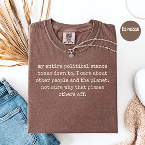 May include: A brown t-shirt with the text "my entire political stance comes down to, I care about other people and the planet. not sure why that pisses others off." A silver necklace is draped across the shirt. The shirt is labeled "EXPRESSO".