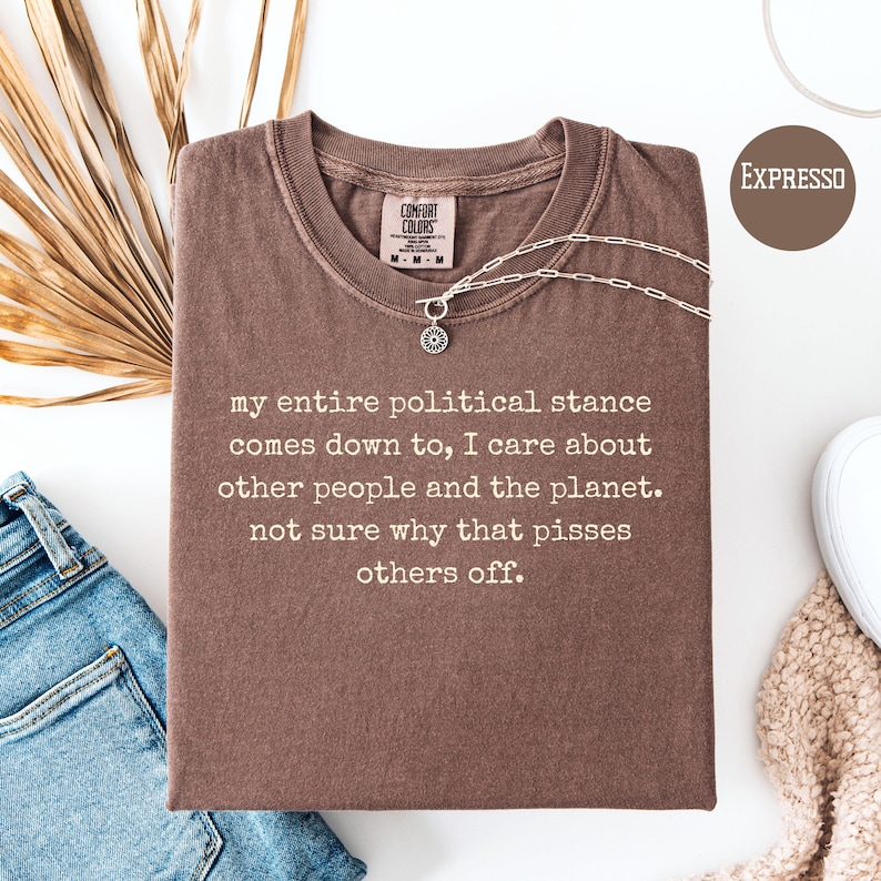 May include: A brown t-shirt with the text "my entire political stance comes down to, I care about other people and the planet. not sure why that pisses others off." A silver necklace is draped across the shirt. The shirt is labeled "EXPRESSO".