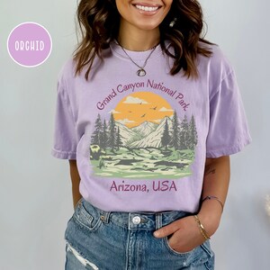 May include: A purple t-shirt with a vintage-style graphic of a mountain landscape with a sunset and the text "Grand Canyon National Park, Arizona, USA".