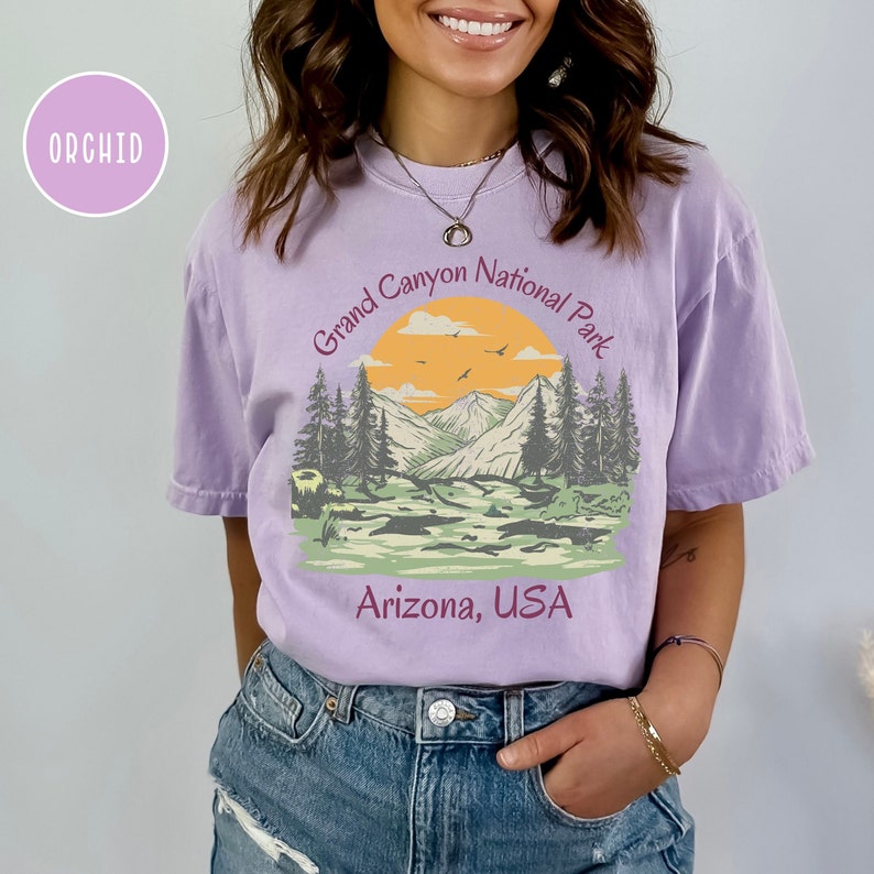 May include: A purple t-shirt with a vintage-style graphic of a mountain landscape with a sunset and the text "Grand Canyon National Park, Arizona, USA".